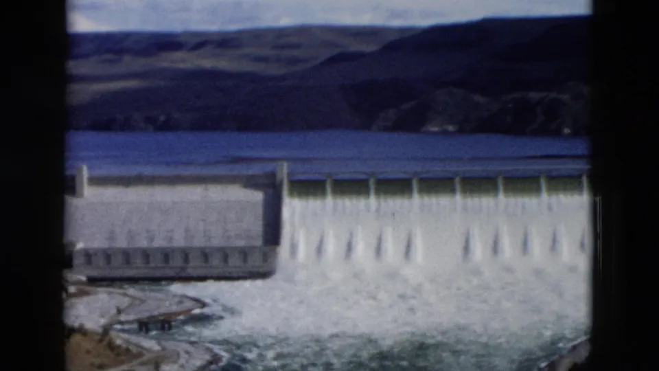 1958 Montana home-movie view of a dam spillway releasing white water, with reservoir and hills in the background.