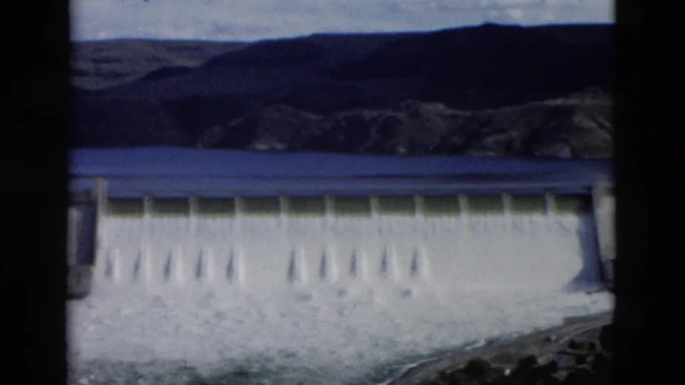 1958 Montana 8mm footage frame showing a broad spillway release, reservoir, hills, and visible film borders.