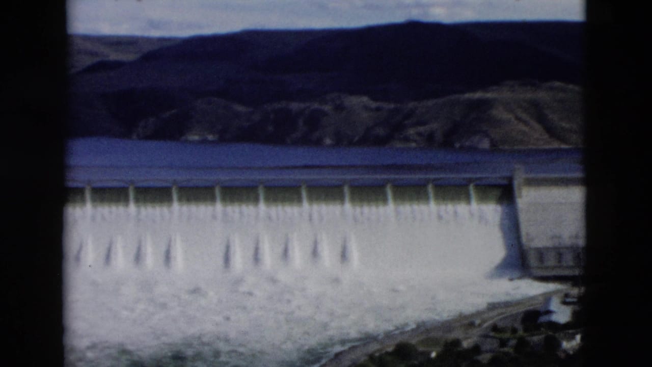 Wide view of a dam releasing water, with a reservoir and dark hills behind it.