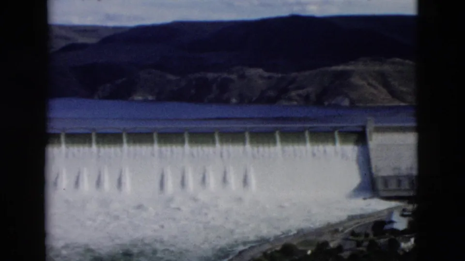 1958 Montana 8mm frame showing a concrete dam spillway with water cascading below a reservoir and rugged hills.