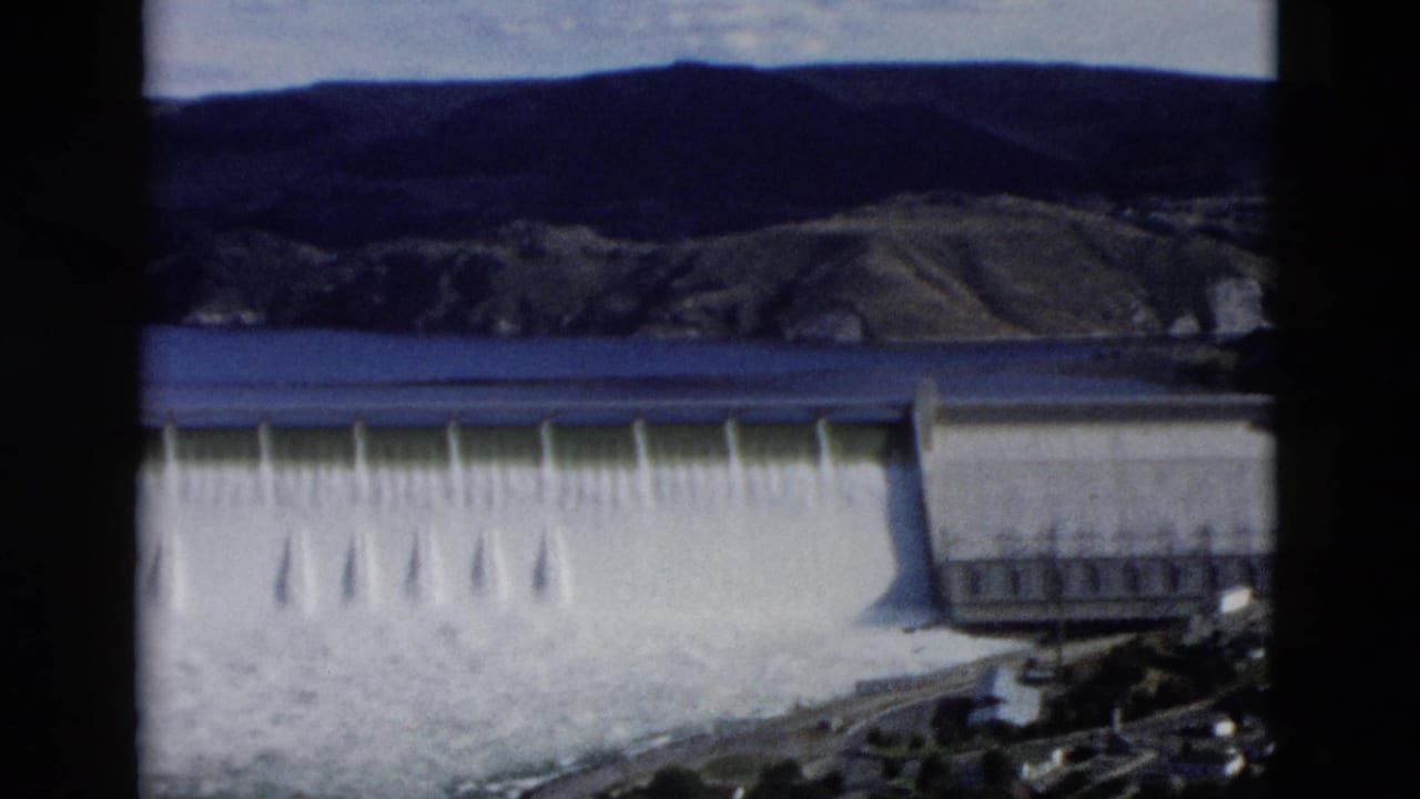 Dam spillway with white water below and hills across the reservoir.