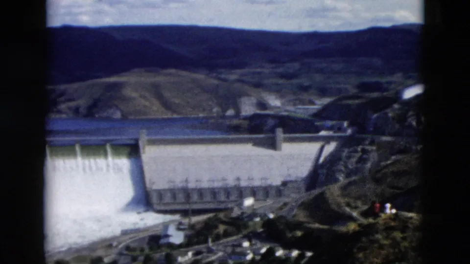 1958 Montana home-movie frame showing the dam, reservoir, hills, and rocky overlook foreground during a water release.