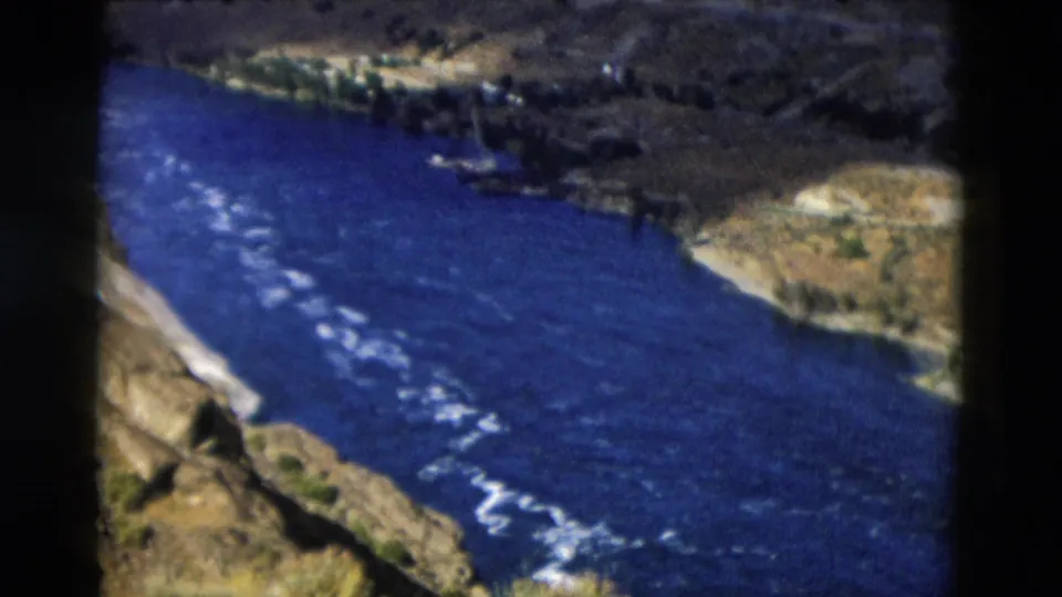 Vintage 8mm Montana view of a blue river flowing through a rugged canyon with steep banks and scattered shoreline vegetation.