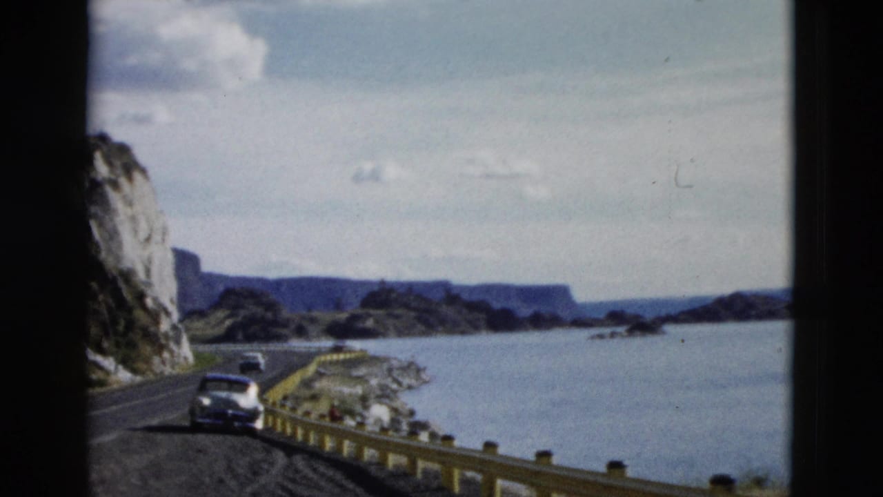 Car on a winding lakeside road beside cliffs and blue water.