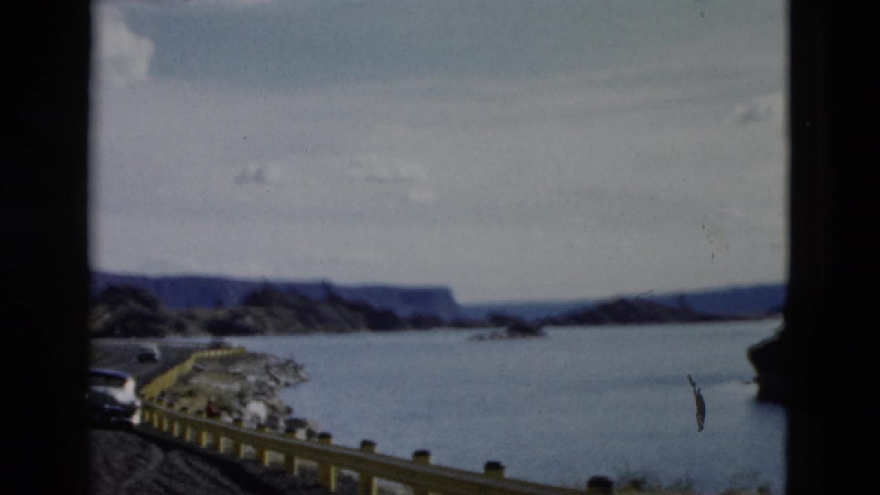 1958 Montana frame with a road edge, yellow guardrail, rocky shoreline, broad water, distant mountains, and cloudy sky.