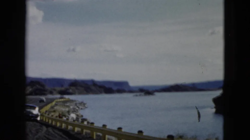 1958 Montana frame with a road edge, yellow guardrail, rocky shoreline, broad water, distant mountains, and cloudy sky.