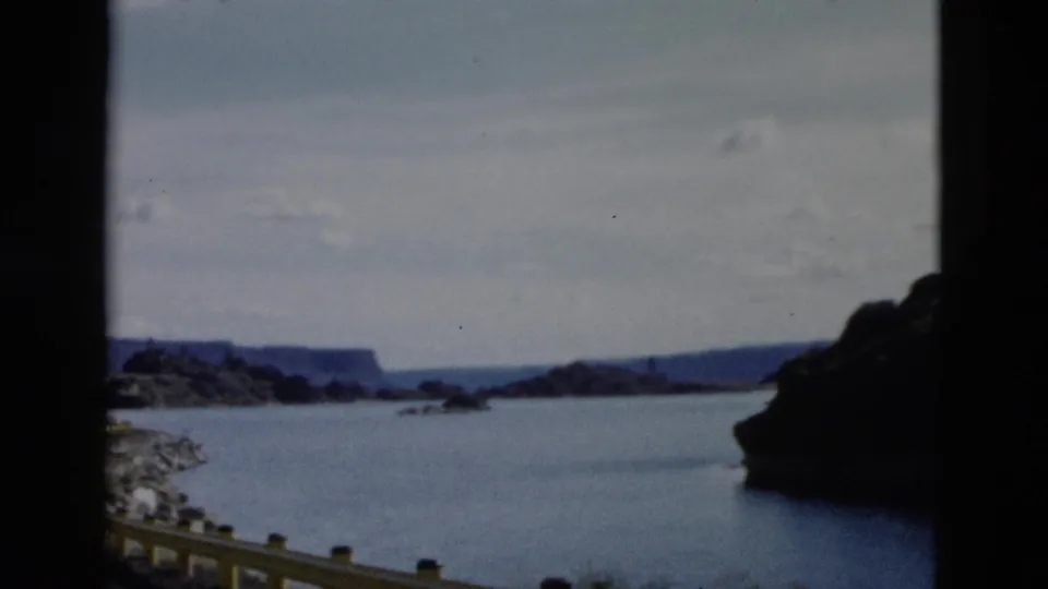 1958 Montana home-movie view across blue water with a yellow guardrail, rocky shoreline, distant ridges, and a dark cliff edge.