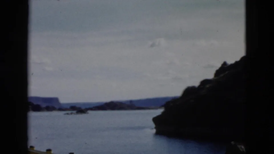 1958 Montana frame looking across a lake or reservoir, framed by dark rock faces, distant mountains, pale sky, and visible 8mm film edges.