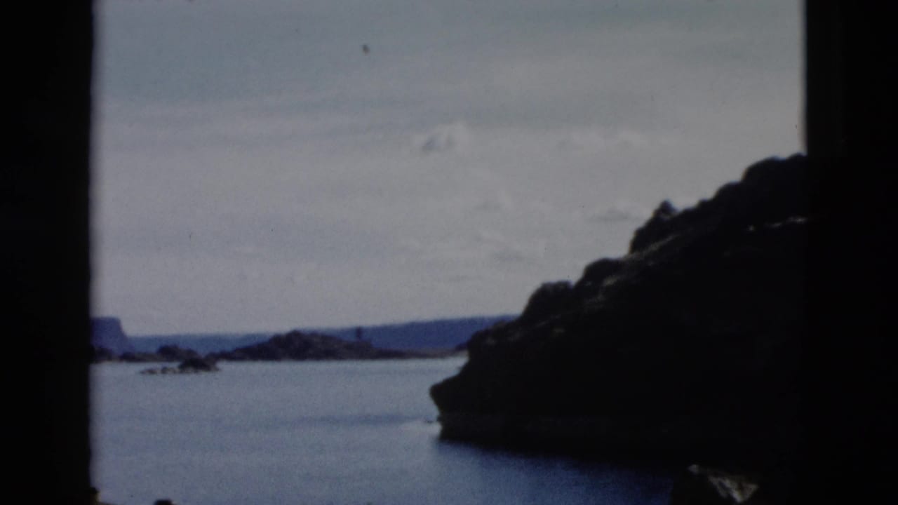 Dark rocky shoreline beside calm blue water under a pale sky, framed by black film edges.