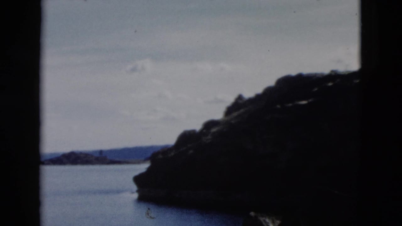 Rocky headland curves into calm water with distant hills and an overcast sky beyond.
