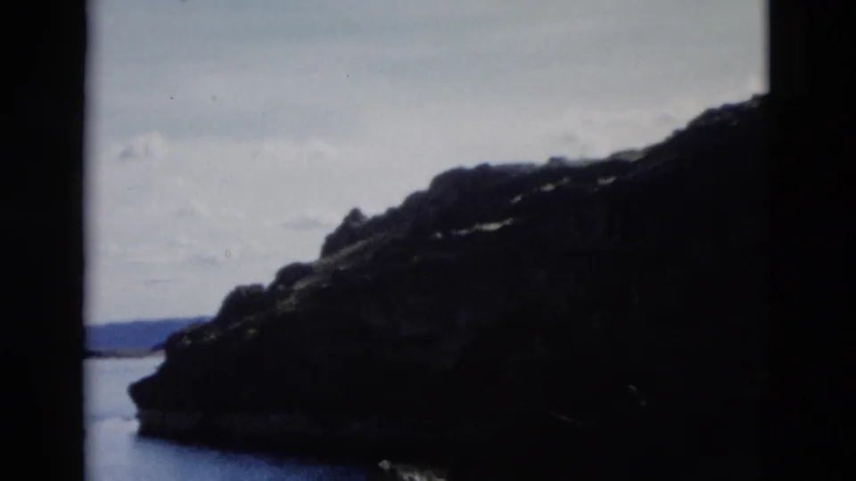 1958 8mm frame showing a dark rocky lakeside or bay slope, calm water, distant hills, and visible film-frame edges.