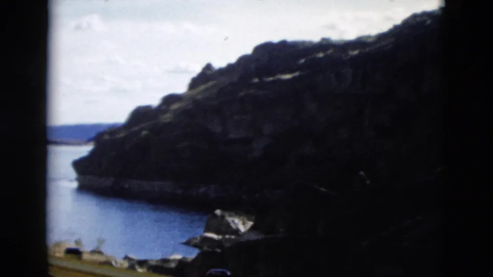 A 1958 Montana 8mm home-movie frame shows a rocky shoreline, quiet water, distant hills, and a small rooflike structure near the bottom edge.