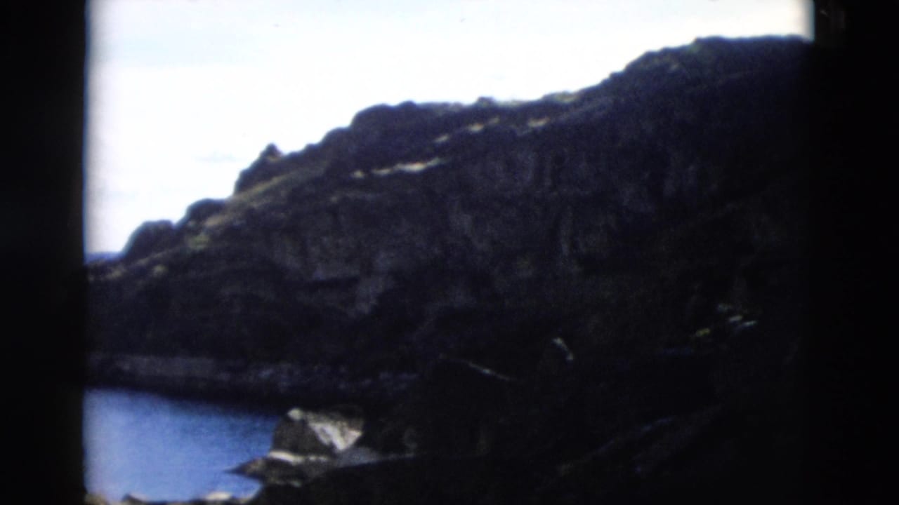 1958 Montana home-movie view of a rocky mountain slope above a body of water, framed with dark film edges.