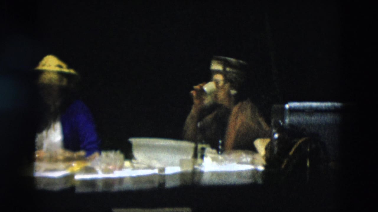 Two women in vintage attire sit at an outdoor dining table, one wearing a conical straw hat and the other lifting a cup.