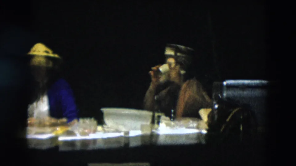 Two women in vintage attire sit at an outdoor dining table, one wearing a conical straw hat and the other lifting a cup.