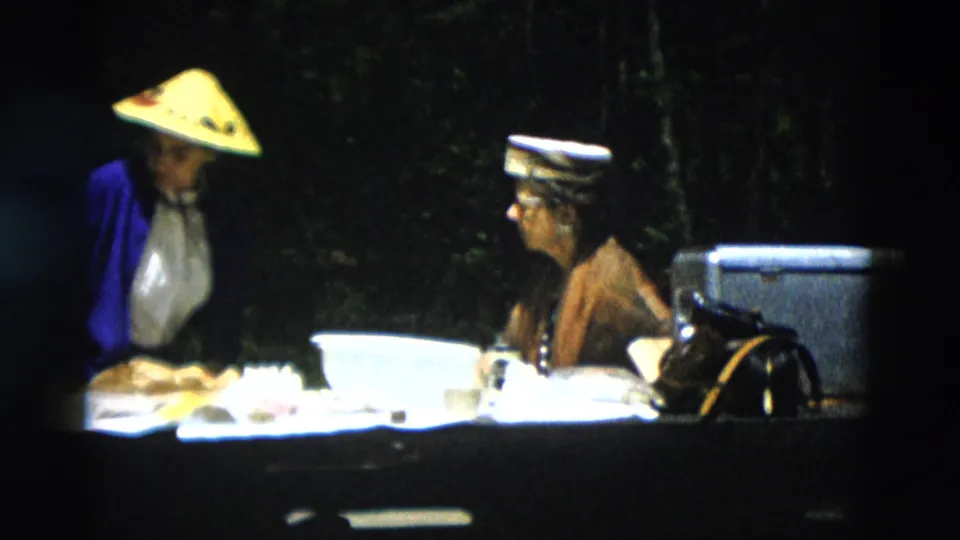 1958 Montana home-movie frame of two women at an outdoor table with food and bowls, including one woman in a straw hat and blue cardigan.