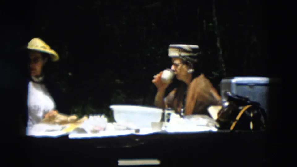Two women continue an outdoor meal, with one drinking from a cup and tableware spread across a white tabletop.