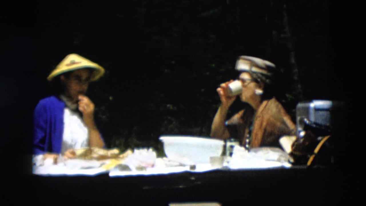 In 1958 Montana home-movie footage, two young women sit at a picnic table, one in a yellow conical hat and one drinking from a cup.