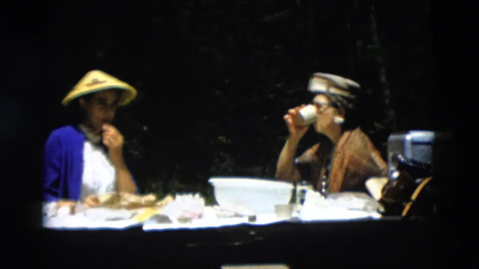 In 1958 Montana home-movie footage, two young women sit at a picnic table, one in a yellow conical hat and one drinking from a cup.