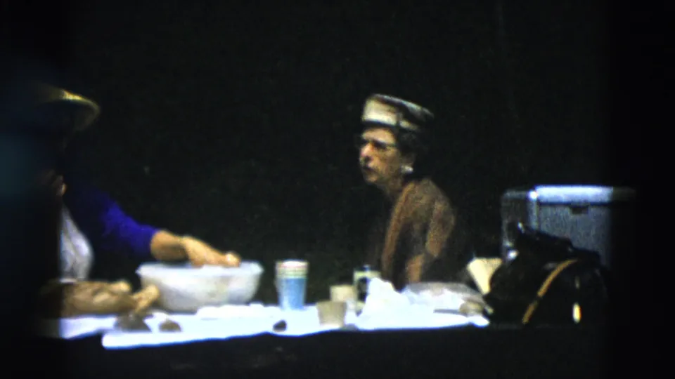 One woman reaches toward the table as the other sits in profile, with cups, bowls, food, a cooler, and bags arranged in the frame.