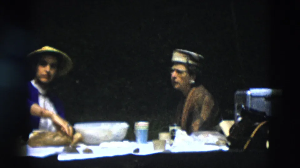 The yellow-hatted woman faces the camera while the other sits in profile beside a cooler, with bread, bowls, and cups on the table.