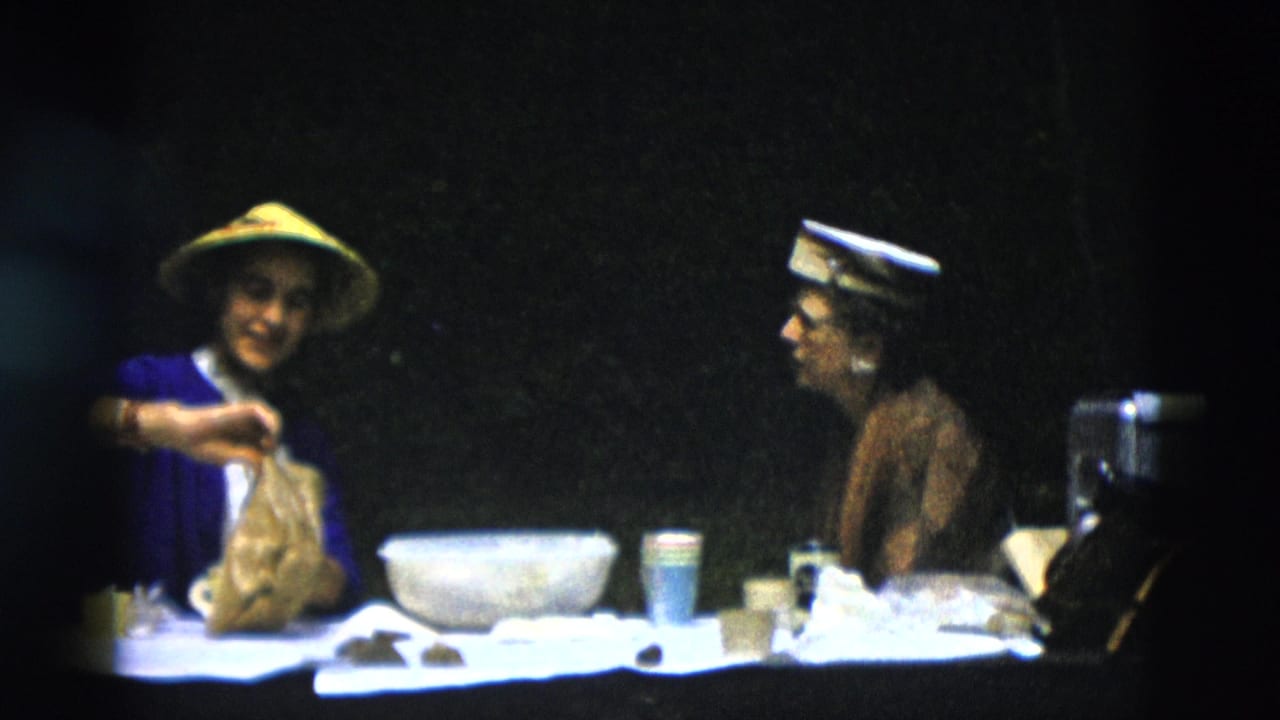 In 1958 Montana home-movie footage, a woman in a blue cardigan and yellow conical hat handles a paper bag at a picnic table while another woman looks on.