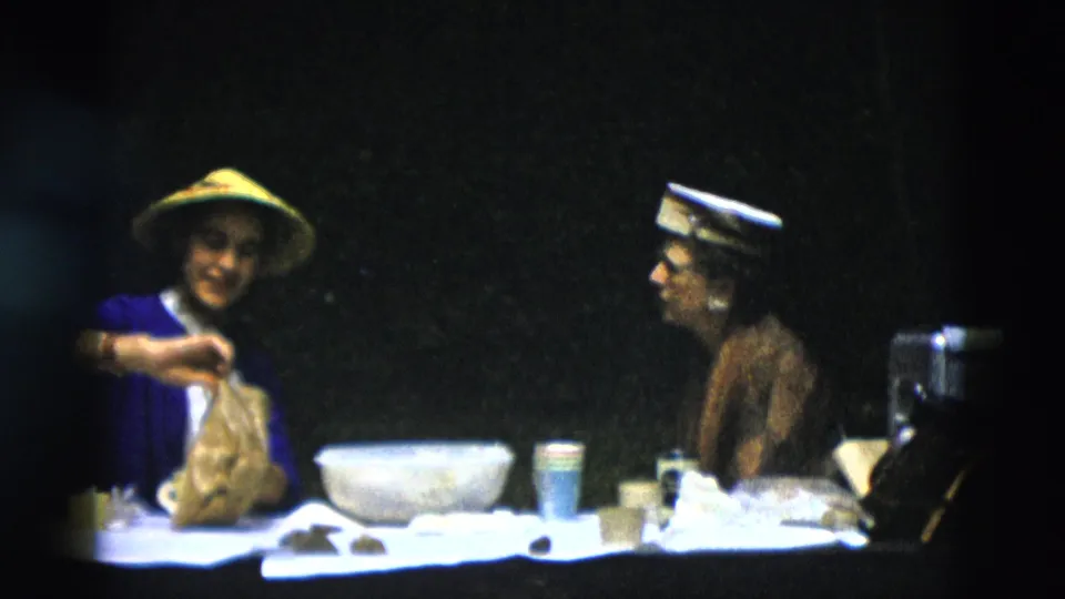 In 1958 Montana home-movie footage, a woman in a blue cardigan and yellow conical hat handles a paper bag at a picnic table while another woman looks on.