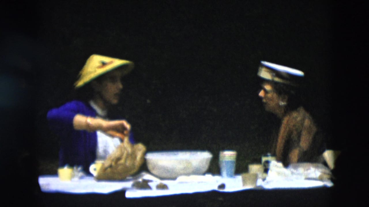 A woman in a yellow conical hat arranges food at a picnic table while another woman sits nearby.