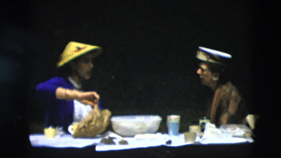 The picnic table scene shows a woman in a blue cardigan and yellow conical hat arranging a food bag beside bowls and cups, with another adult seated at right.