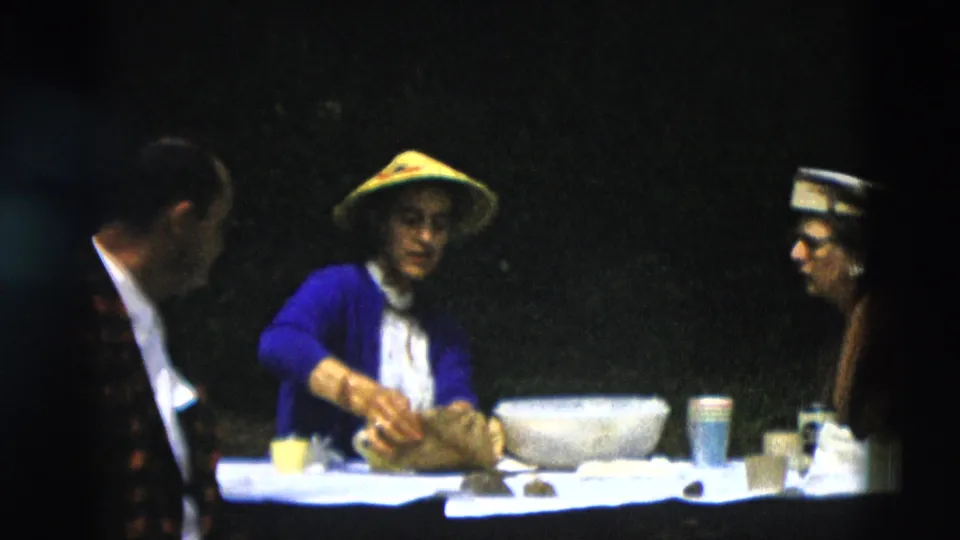 A man in a patterned jacket sits at the edge of the frame while a woman in a yellow conical hat works with food on a picnic table in Montana home-movie footage.