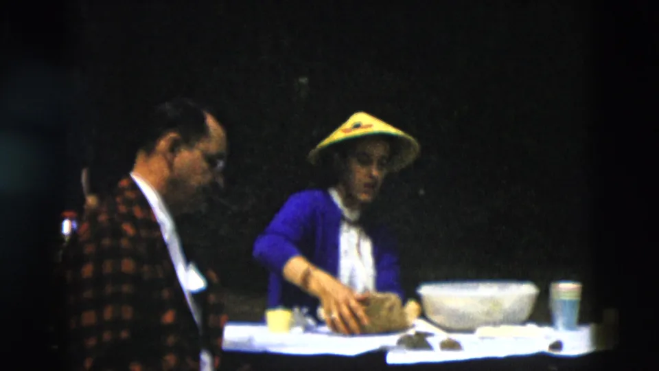 Adults remain gathered around the picnic table as the woman in the yellow conical hat rests her hand on a food bag beside a large bowl and cups.