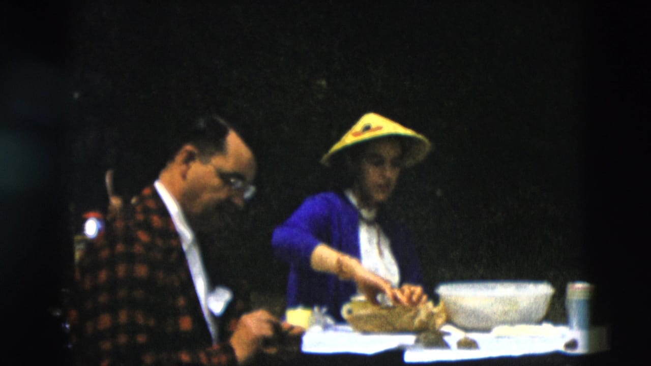 Adults seated at a table as a woman in a yellow conical hat reaches toward food.