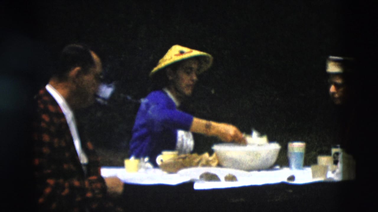 Woman in blue reaches into a large bowl while a seated man smokes nearby.