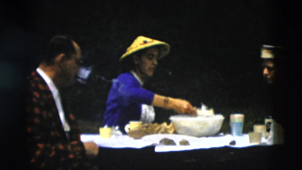 1958 Montana home-movie frame with a woman in a yellow hat preparing food beside a seated man and tableware.