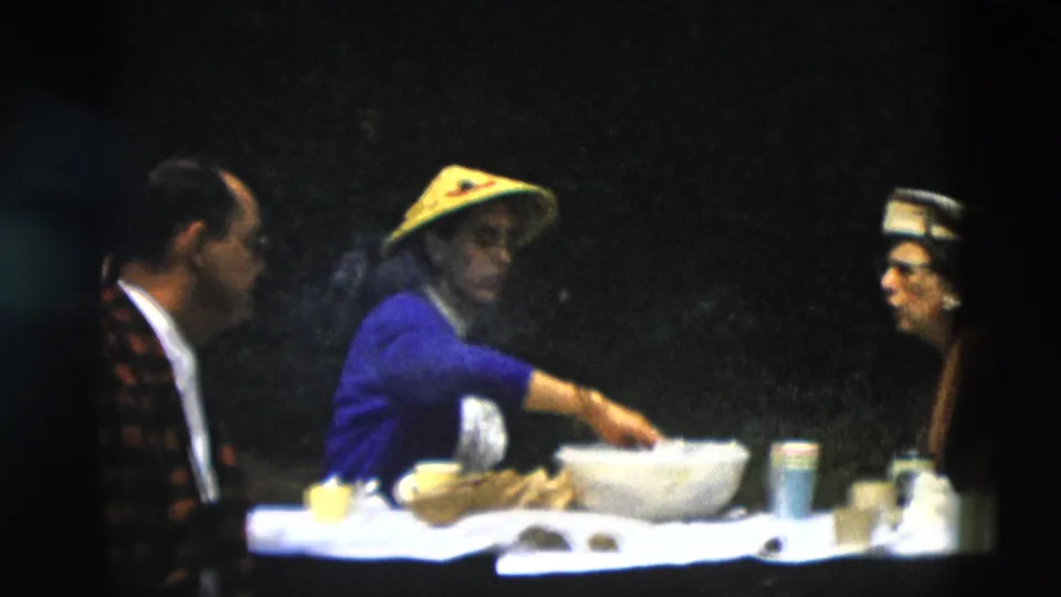 1958 Montana 8mm still showing three adults at a table, with a woman in a yellow hat reaching into a bowl.