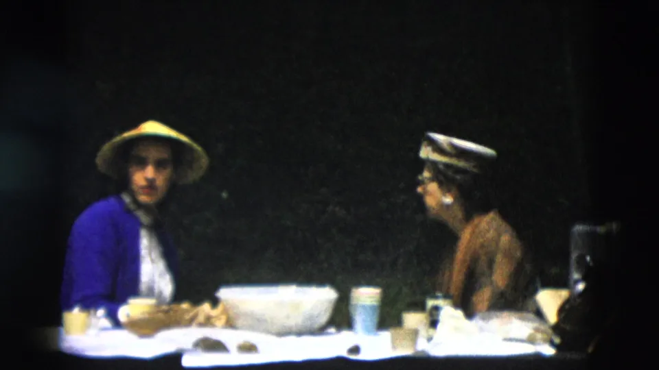 1958 Montana home-movie frame of two women seated across a table, one wearing a yellow conical hat and blue cardigan.
