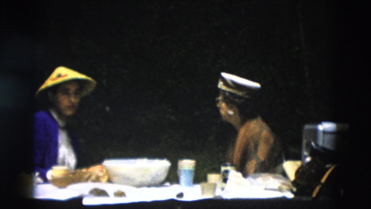Two adults sit at an outdoor table with cups and bowls, one wearing a conical straw hat.