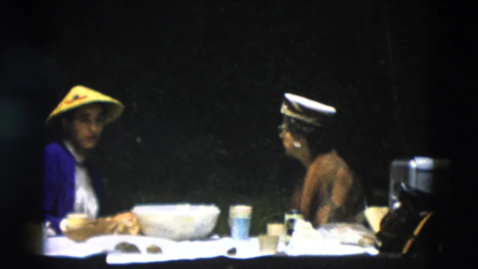 1958 Montana home-movie frame showing two adults in conversation at an outdoor table, with one wearing a conical straw hat and blue jacket.
