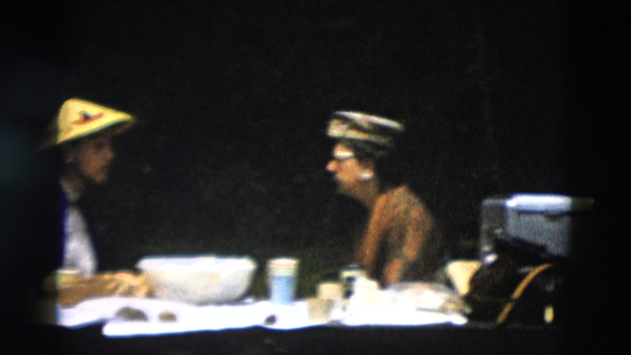 Two adults appear to talk across a picnic-style table in a 1958 Montana travel scene, with tableware and dark foliage in the background.