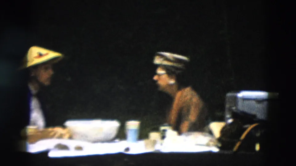 Two adults appear to talk across a picnic-style table in a 1958 Montana travel scene, with tableware and dark foliage in the background.