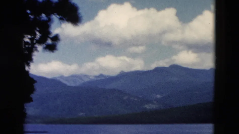 1958 Montana scenery frame with a lake, distant mountain ridges, cloudy sky, and dark tree silhouettes at the edges of the 8mm frame.