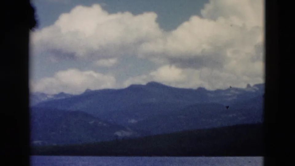 A 1958 Montana home-movie landscape view shows a broad lake, layered mountain ridges, and a bright sky filled with clouds.