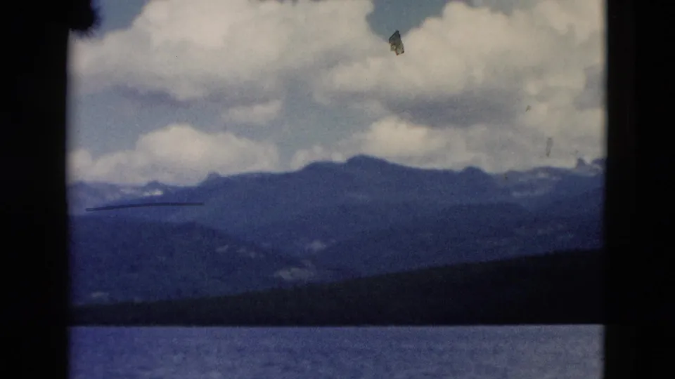 1958 Yellowstone home-movie frame showing a lake foreground, dark shoreline, distant mountain ridges, and large clouds.