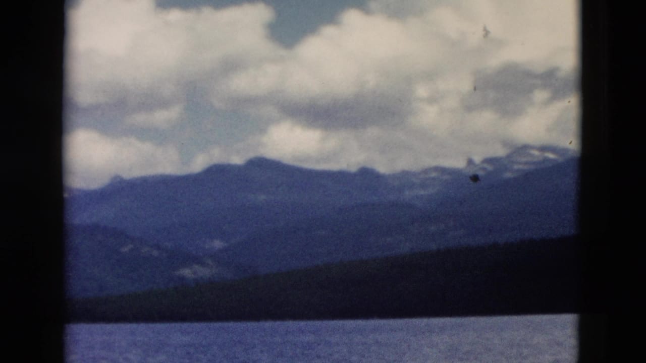 1958 Yellowstone frame with lake water, a shadowed tree line, blue mountain silhouettes, and bright clouds overhead.