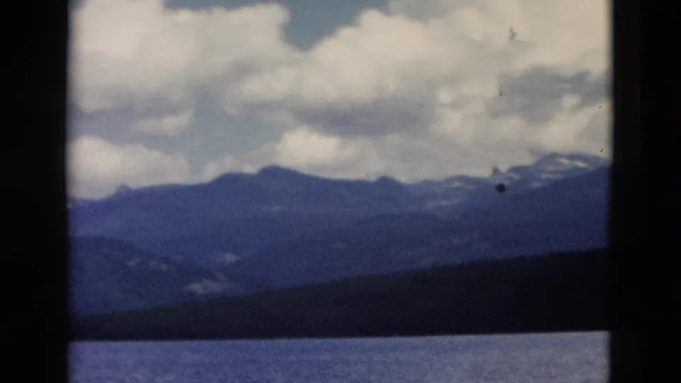 1958 Yellowstone frame with lake water, a shadowed tree line, blue mountain silhouettes, and bright clouds overhead.