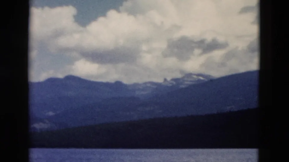 1958 Yellowstone home-movie still with a soft view across lake water to forest, blue ridges, and cloud cover.