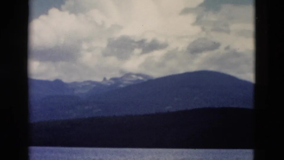 1958 Yellowstone frame showing lake foreground, wooded shore, rounded mountain slope, snow flecks, and drifting clouds.