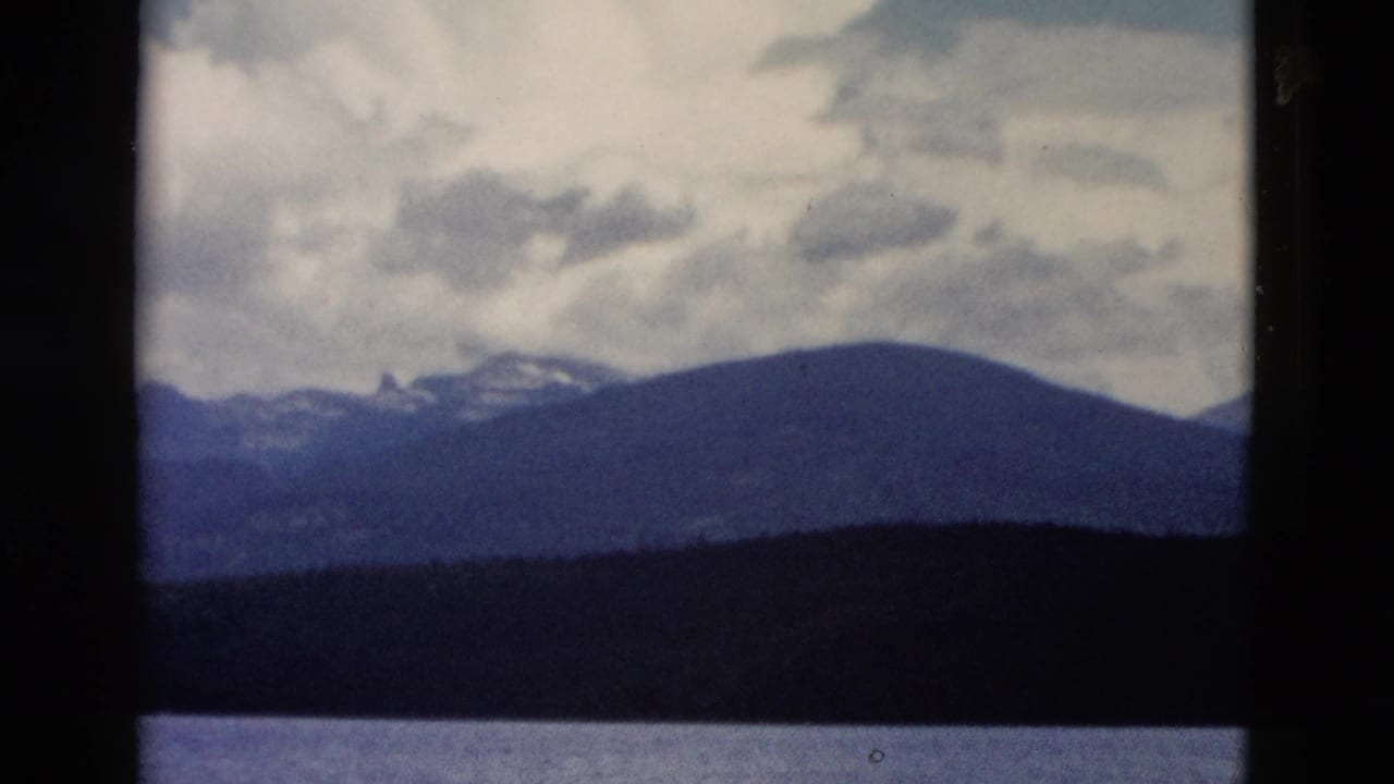 Distant mountains beyond calm water under a cloudy sky, framed by dark film borders.