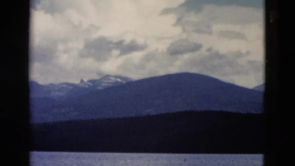 1958 Yellowstone home-movie frame showing calm water, a dark shoreline, and distant mountain ridges beneath clouds.