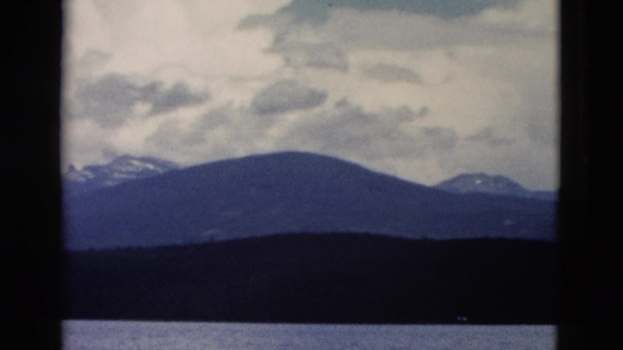 1958 Yellowstone landscape frame with blue-gray mountain ridges, still water, and overcast sky in a soft 8mm scan.